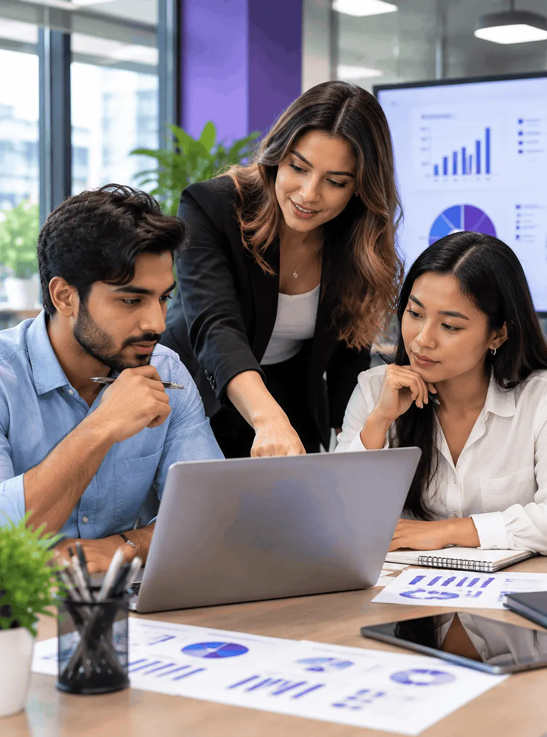 Three professionals discussing project reports using a laptop and tablet in a modern office.